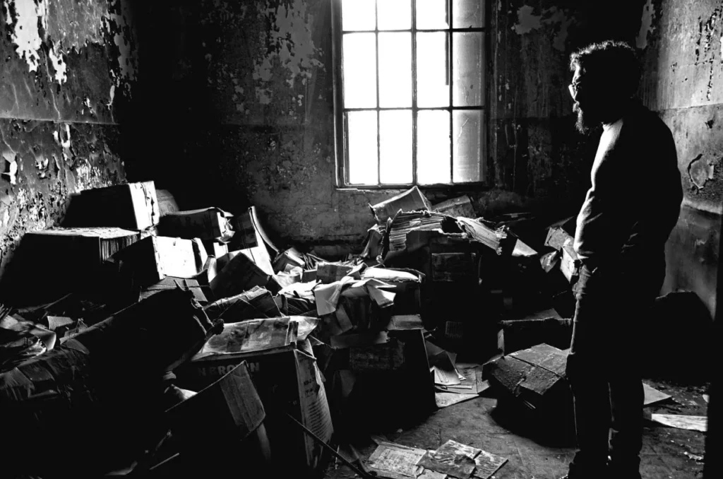 un escritor de pie en su propio habitat, o sea una habitación llena de periódicos, libros y fotos tiradas al suelo